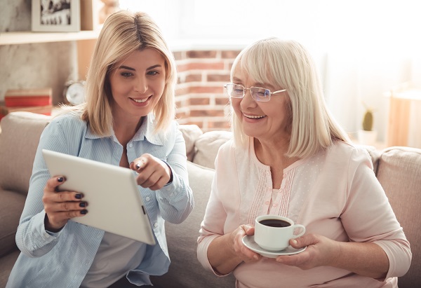 Beautiful mature mother and her adult daughter are drinking coffee, using a digital tablet and smiling while sitting on couch at home